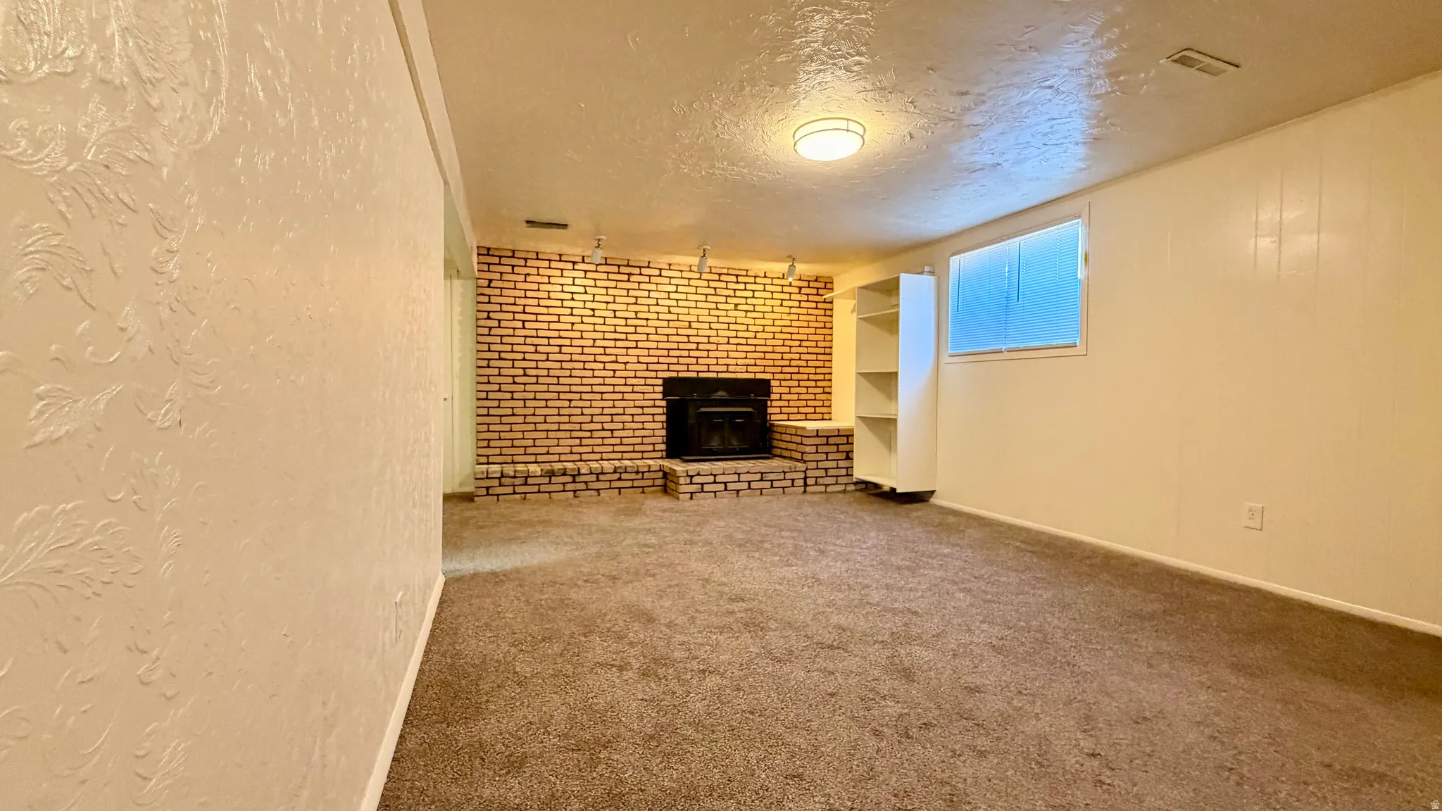 Carpeted room featuring a brick fireplace with a raised hearth, built-in shelving, a window, and overhead lighting
