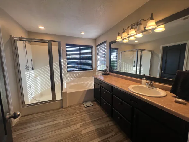 Bathroom featuring a glass-enclosed shower, built-in soaking tub, wood-finish flooring, a double vanity with an integrated sink, and an expansive mirror