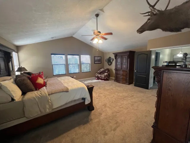 Vaulted ceiling with a ceiling fan, plush carpeting, multiple windows providing natural light, and an integrated door leading to an additional space