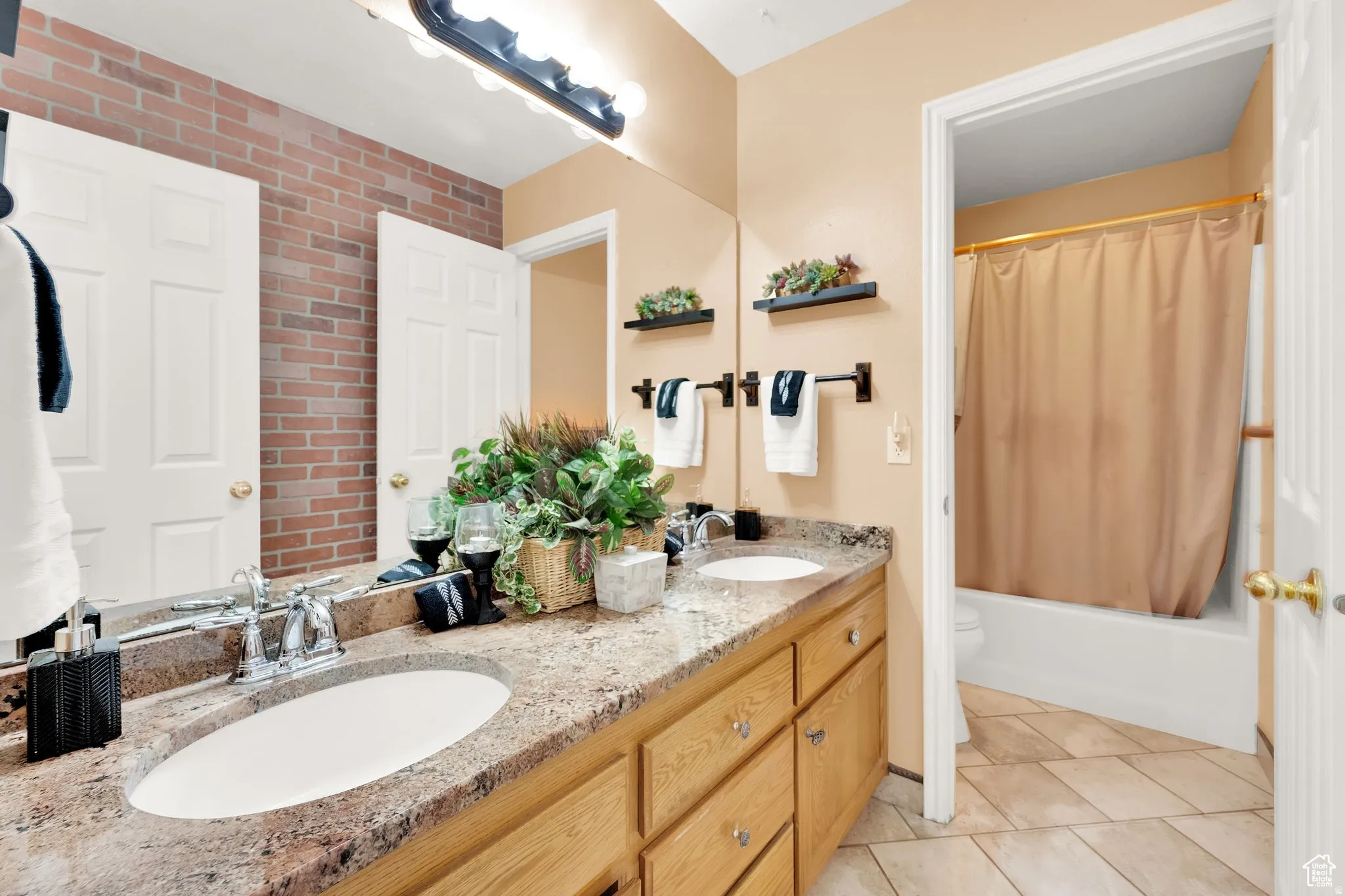 Dual under-mount sinks set into a speckled countertop, light wood-finish cabinetry, and a brick-patterned accent wall