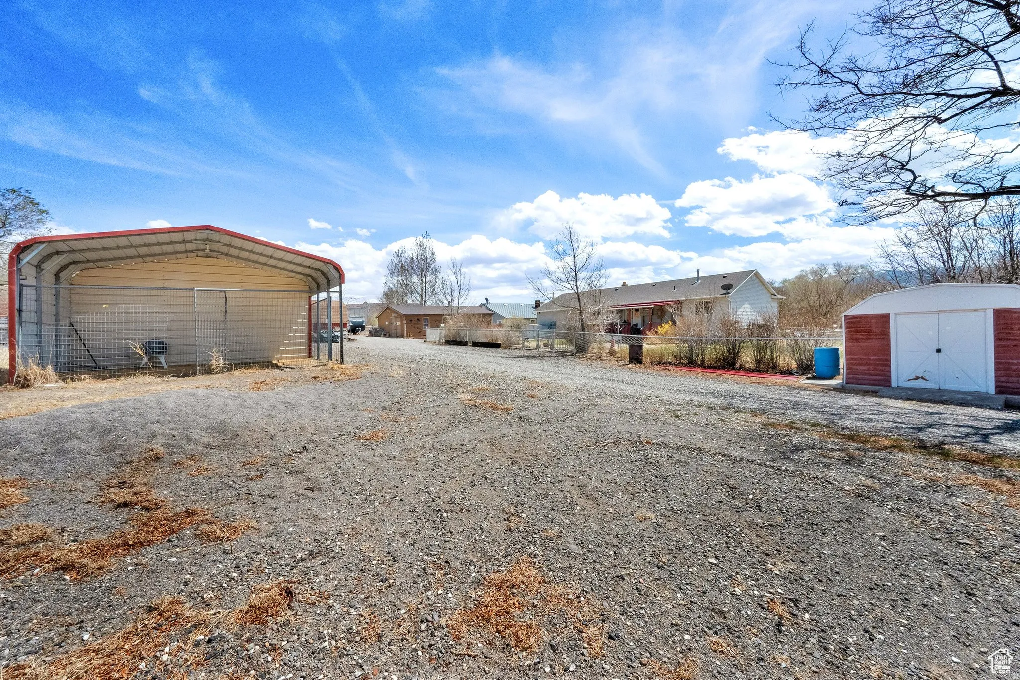 Expansive gravel lot featuring a metal carport structure with a red roof, a separate storage shed with white doors and red siding, and a fenced perimeter