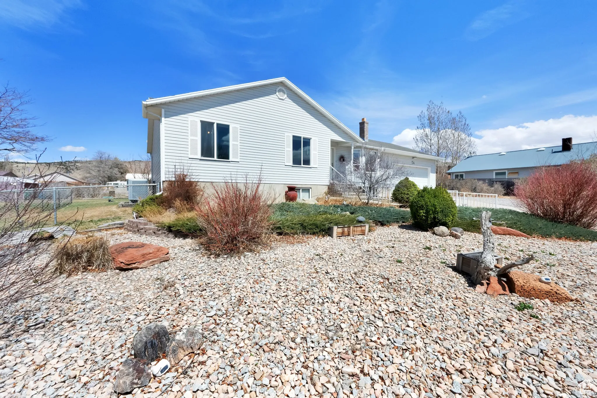 White vinyl siding exterior with an attached garage, featuring a gravel front yard, established shrubs, and decorative rockwork