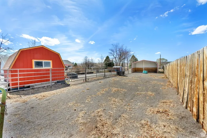 Gravel yard featuring a red barn-style shed with white window trim, metal fencing, a covered carport, and a rustic wood fence