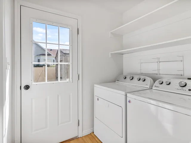 Dedicated laundry room featuring side-by-side washer and dryer units with control panels, open shelving for storage, wood-finish flooring, and natural light from an exterior door with multiple panes and access to outdoor space