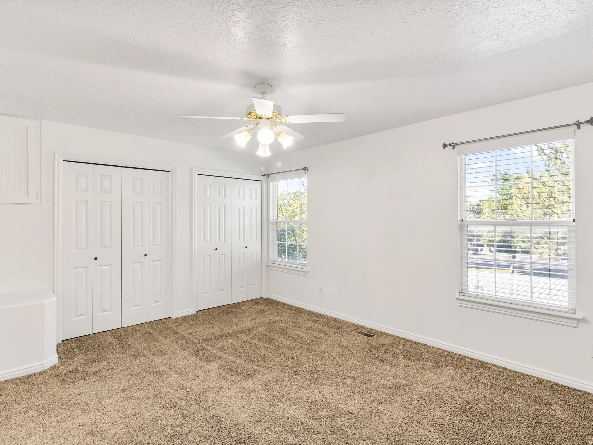 Room featuring dual bi-fold door closets, two windows with blinds, a ceiling fan with light fixture, and neutral carpet flooring