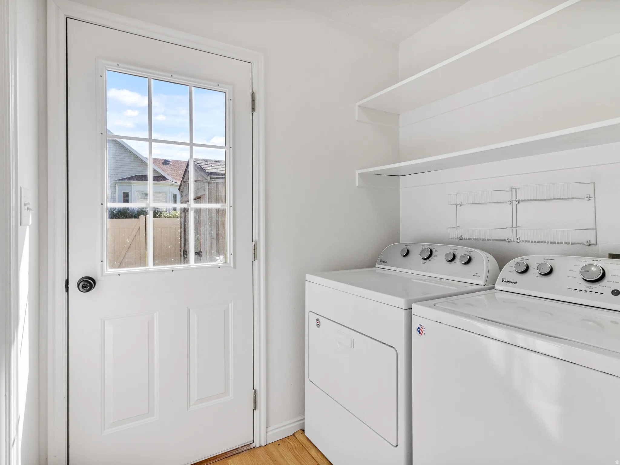 Utility room featuring a white paneled door with glass inserts, wood-finish flooring, two white wall-mounted shelves, and additional wire shelving