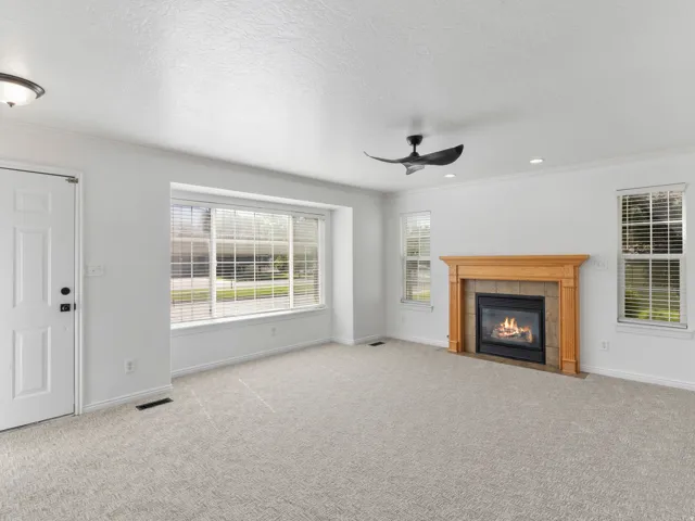 Spacious living area featuring neutral carpet flooring, a wood-framed fireplace with gas insert, expansive multi-pane windows with plantation shutters, ceiling fan, and glass door entry with sidelights