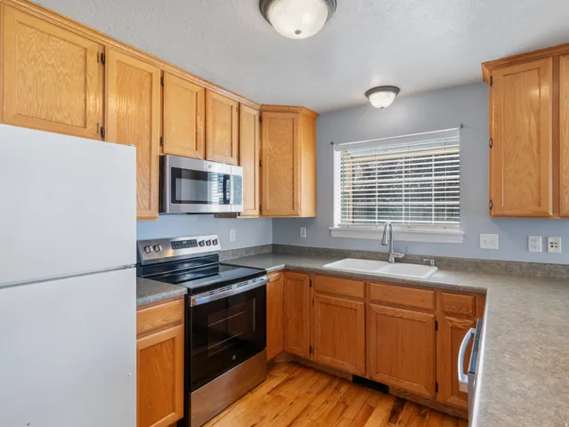 Efficient kitchen featuring warm wood cabinetry, stainless steel appliances including a built-in microwave, electric range, and refrigerator, laminate countertops, undermount sink with chrome faucet, and wood-finish flooring throughout