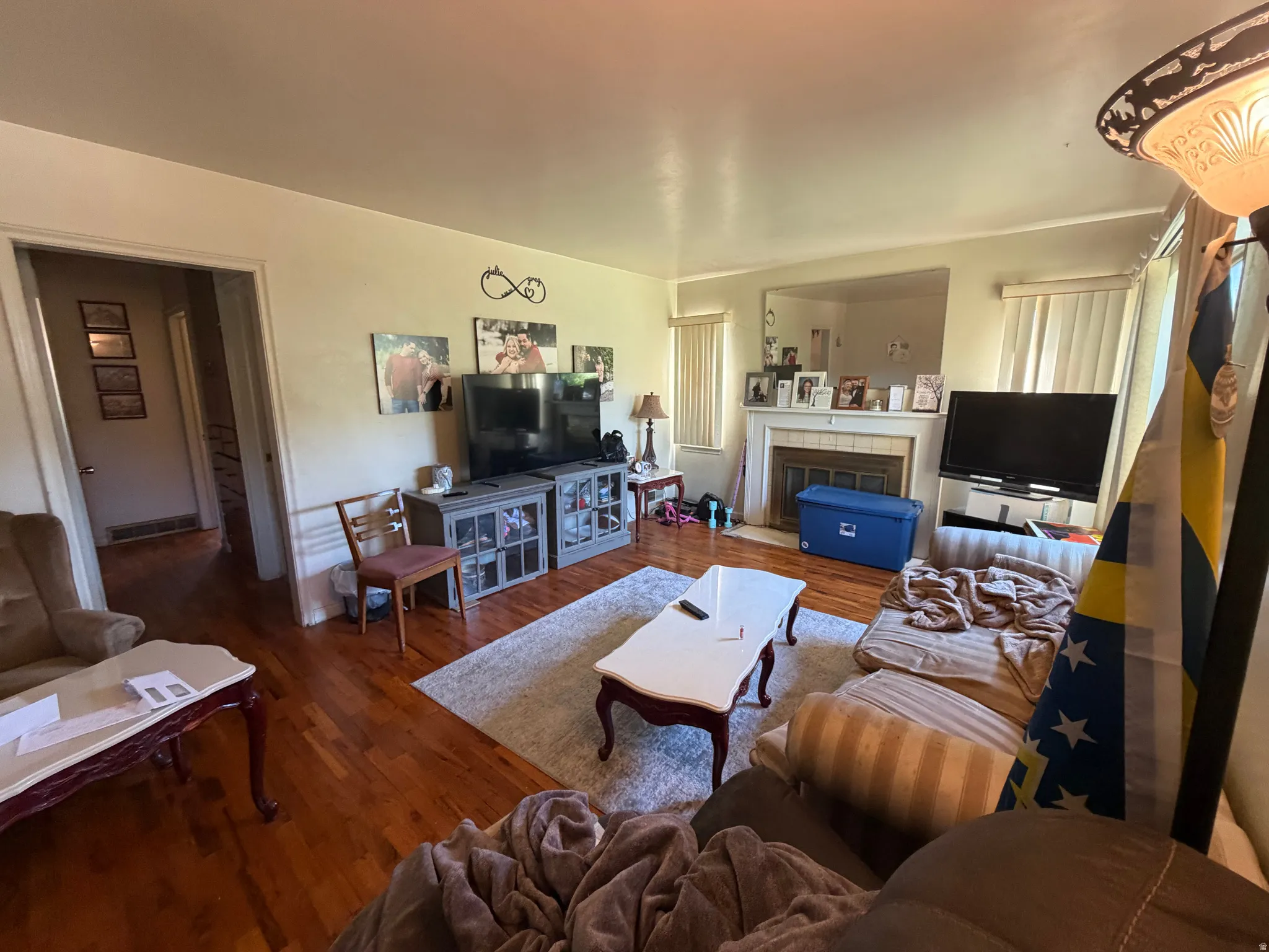 Living area featuring wood-finish flooring, a white mantelpiece, built-in fireplace, and multiple windows with vertical blinds