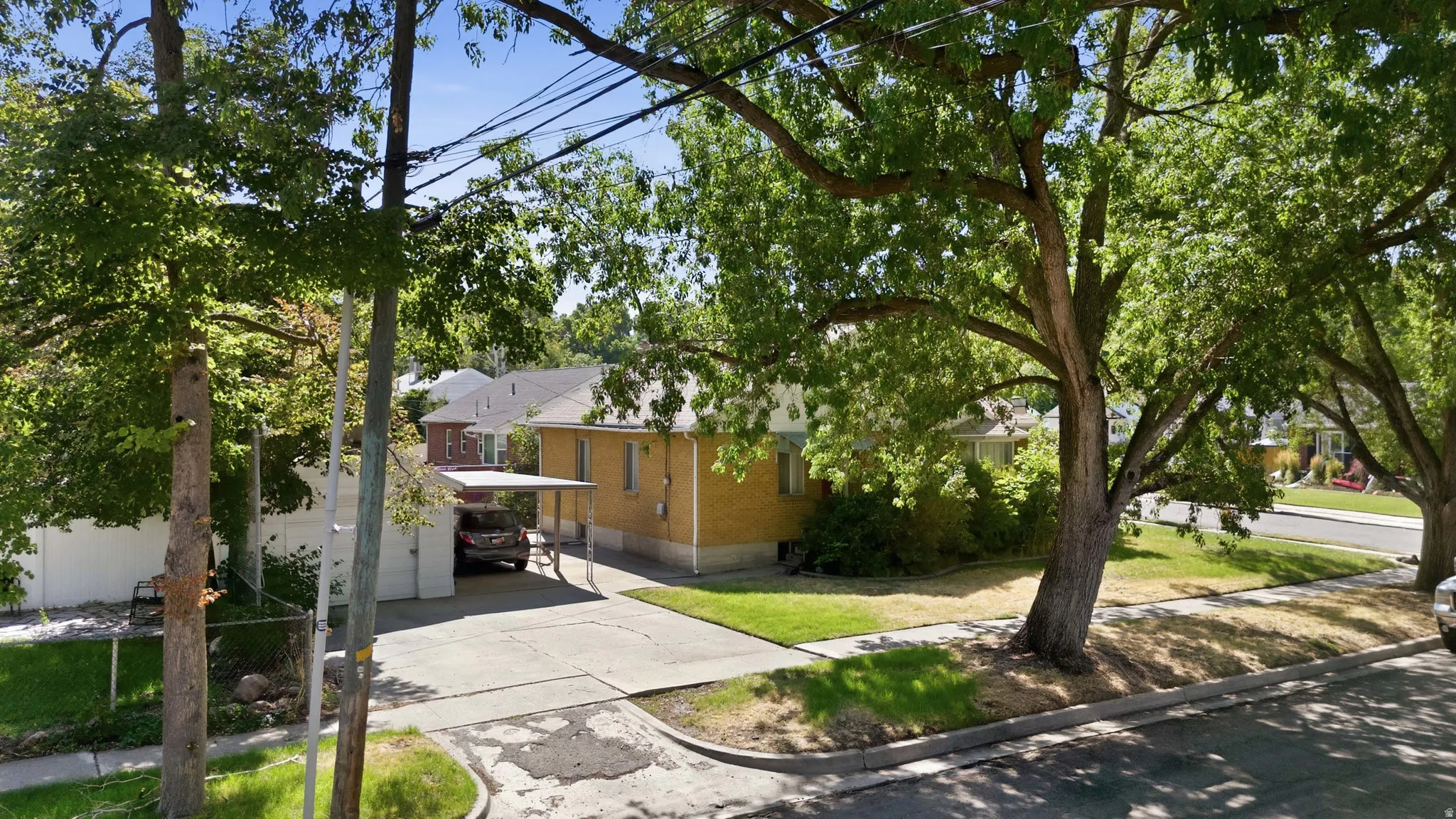 Yellow brick exterior with a covered carport, mature shade trees, and a concrete driveway