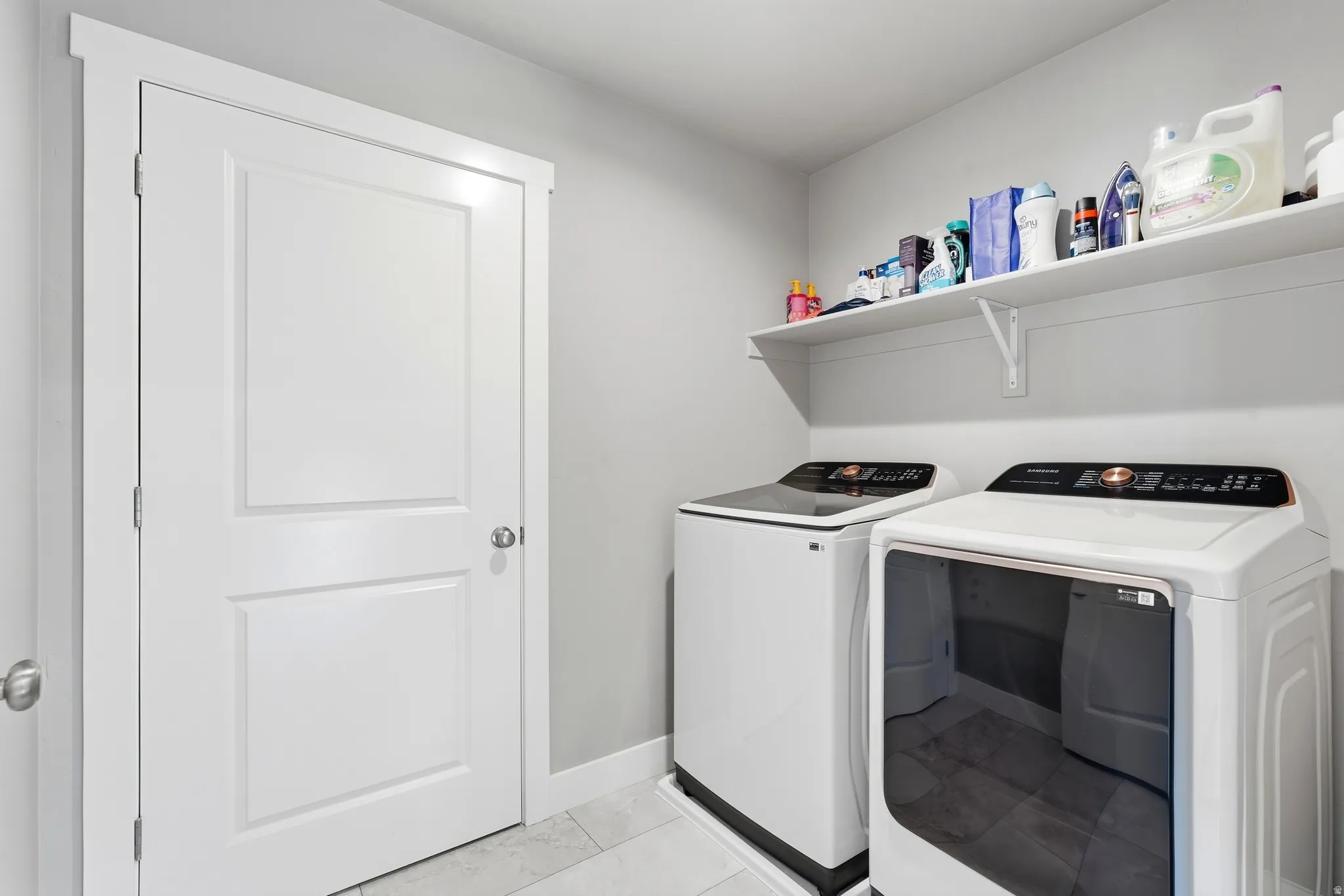 Dedicated laundry area featuring a white paneled door, light gray wall paint, tiled flooring, and a long white floating shelf
