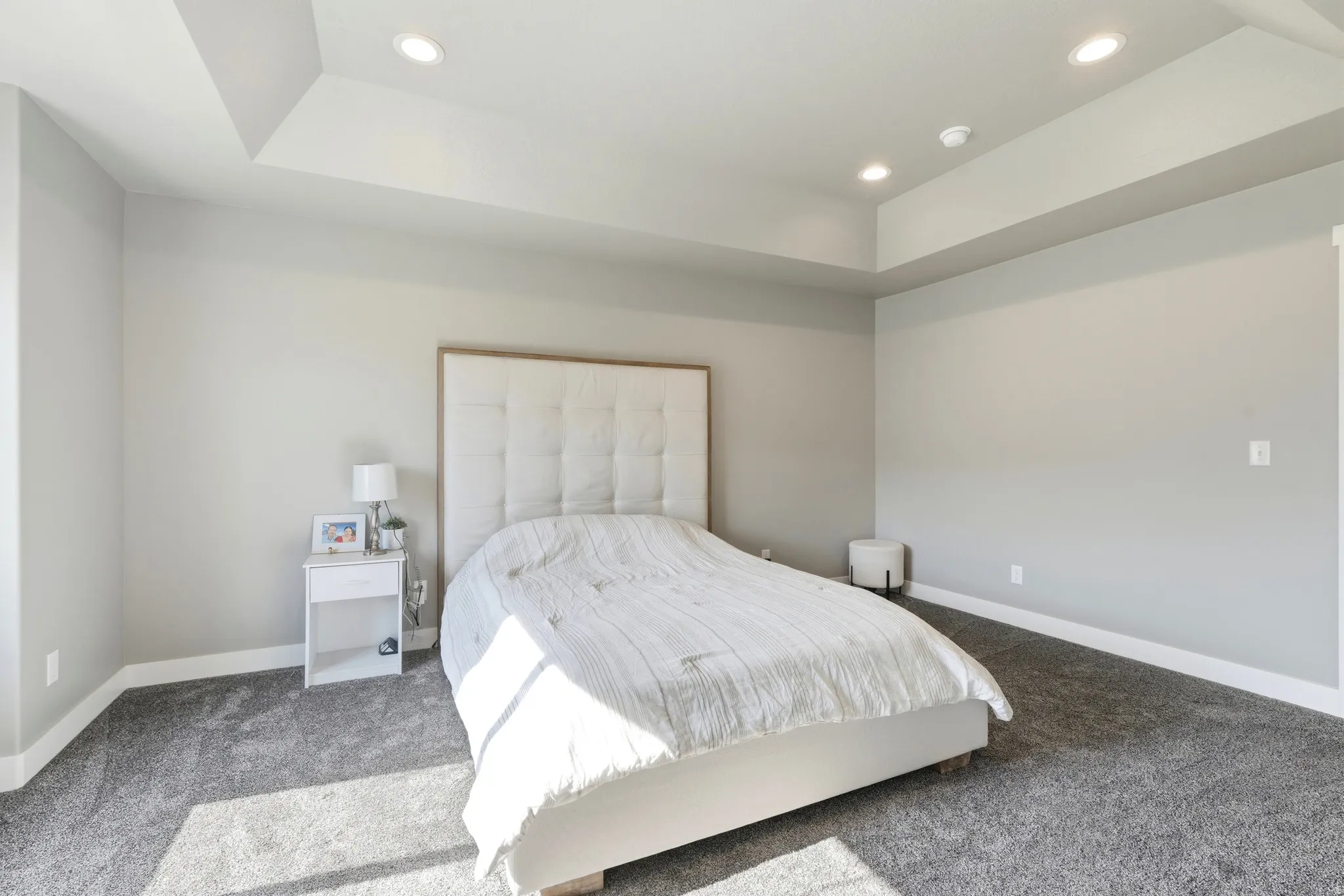 Recessed lighting illuminates a room featuring a tray ceiling, light gray wall paint, white baseboards, and gray carpeting