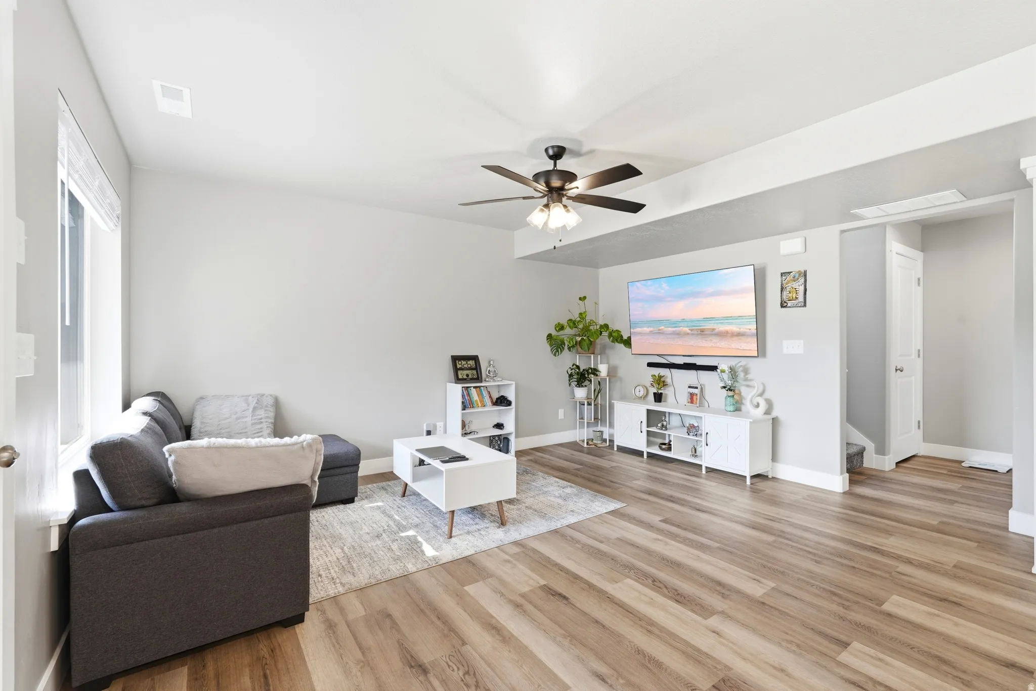 Spacious living area featuring wood-finish flooring, light gray walls, a ceiling fan with integrated lighting, a large window with blinds, and white baseboards
