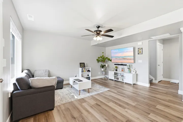 Spacious living area featuring wood-finish flooring, light gray walls, a ceiling fan with integrated lighting, a large window with blinds, and white baseboards