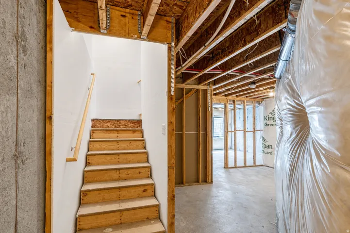 Unfinished basement with exposed wood framing, concrete flooring, and an unfinished staircase featuring a wood handrail