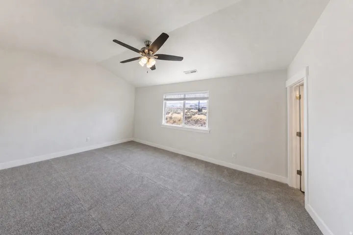 Spacious room featuring vaulted ceilings, a ceiling fan with integrated lighting, textured gray carpet, and a large window with a landscape backdrop