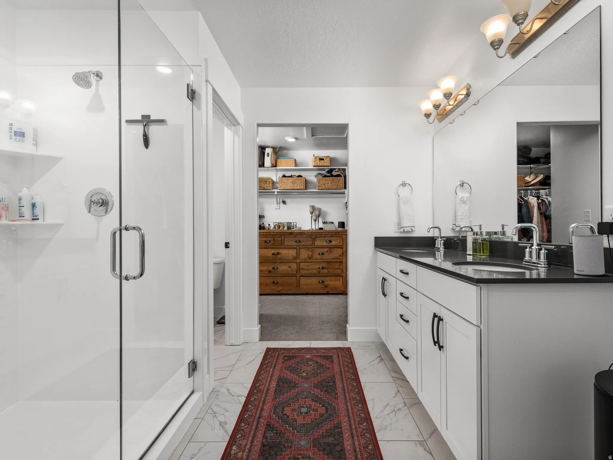 Bathroom featuring a double vanity with dark countertops and dual sinks, a large mirror, and glass-enclosed shower with built-in shelving