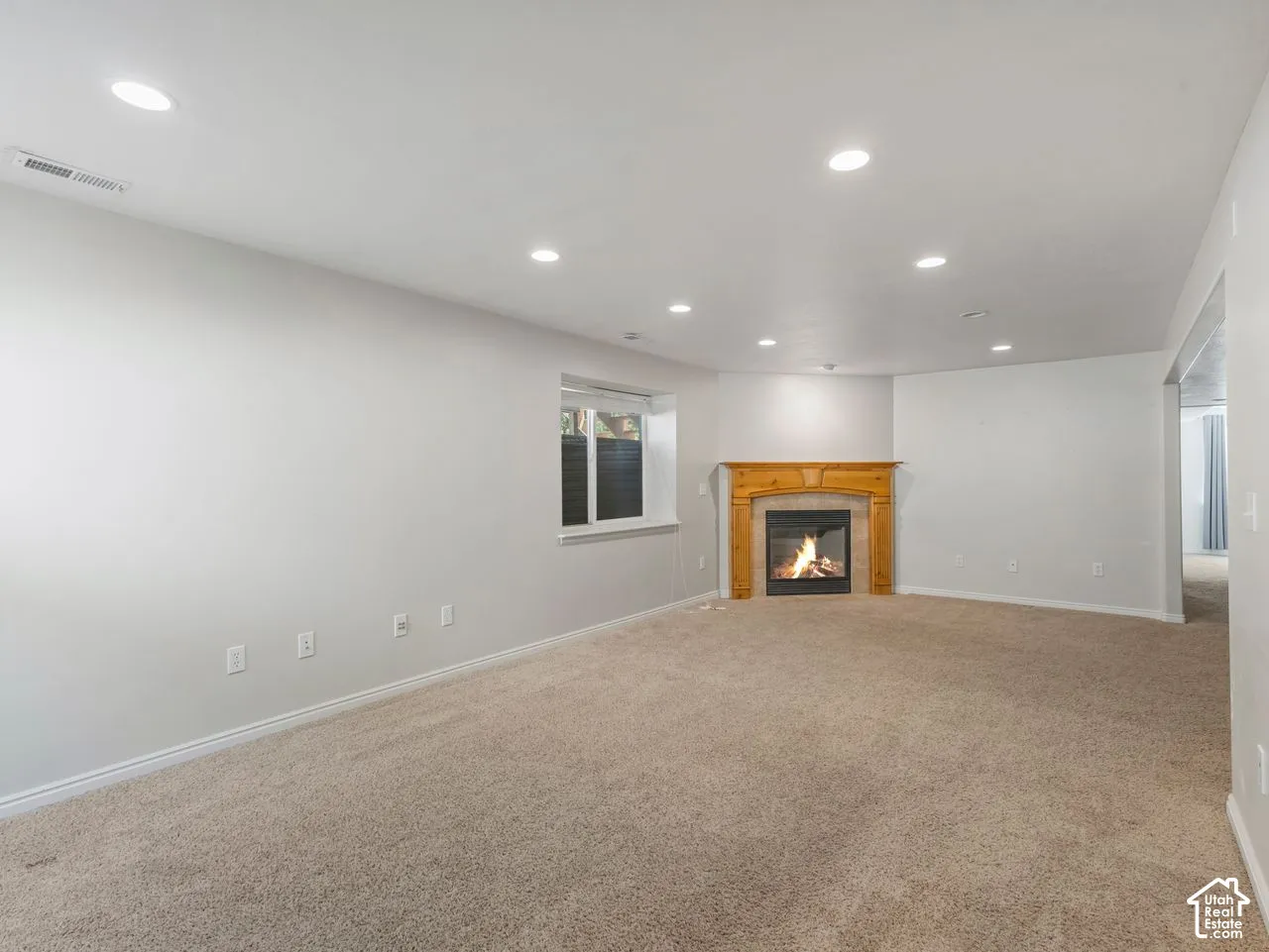 Corner fireplace with a light wood mantel, recessed lighting, neutral wall paint, and a window with a white frame