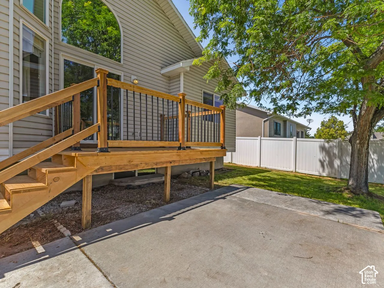 Wood deck with black metal balusters, concrete patio, lawn area, and white privacy fence