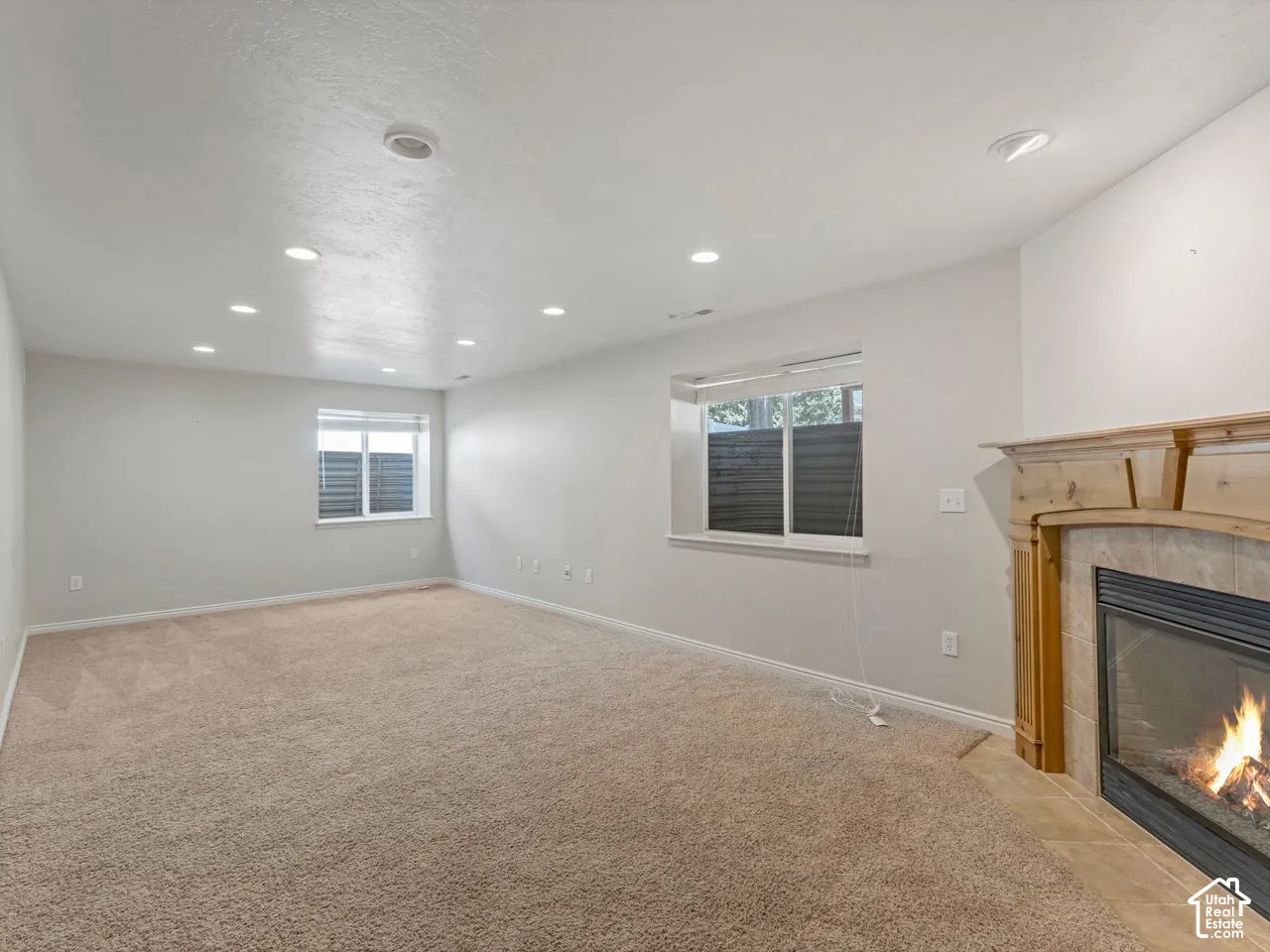 Spacious room featuring a fireplace with a light wood mantel, recessed lighting, two windows with blinds, and carpeting throughout