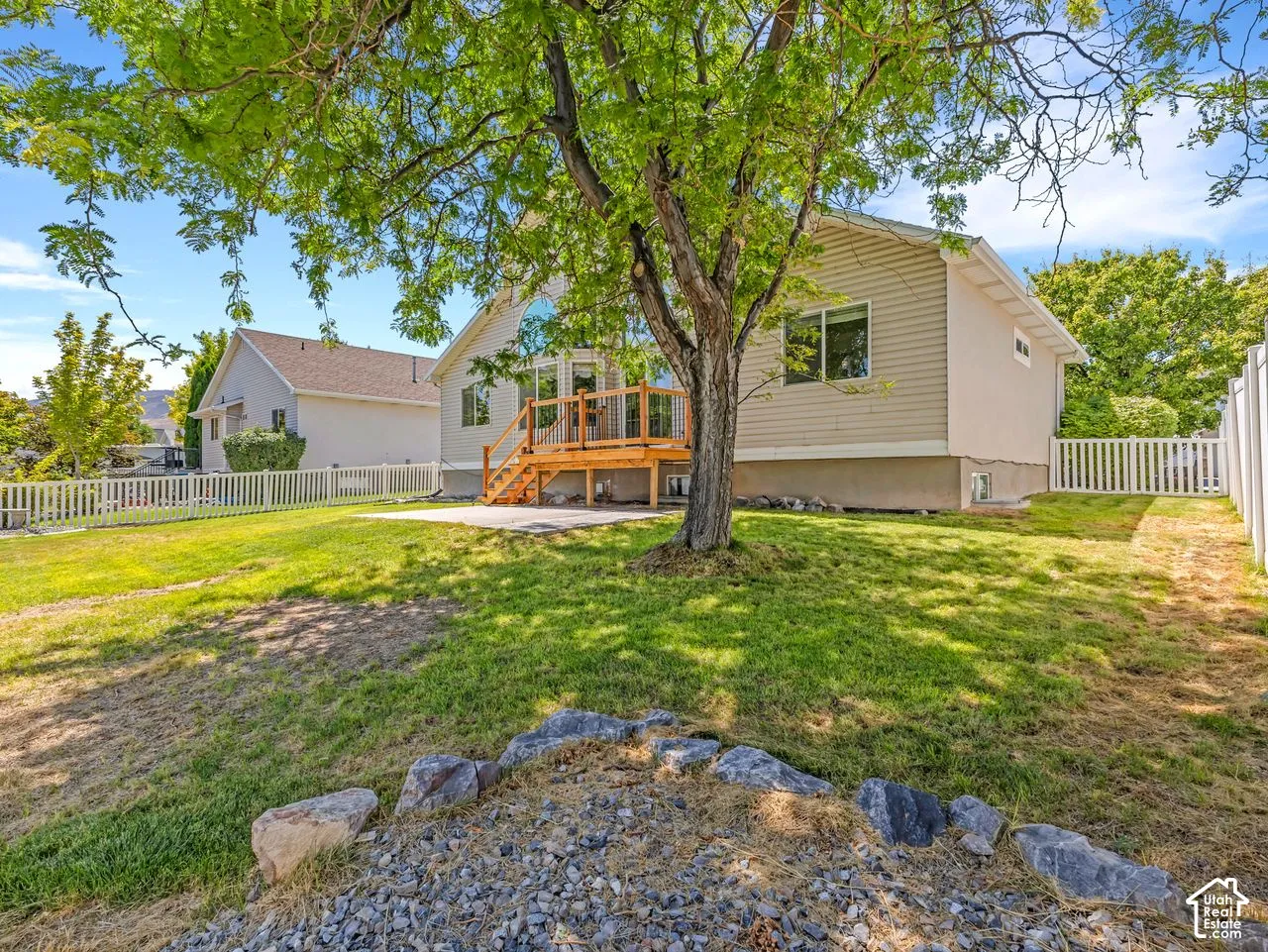 Rear exterior featuring a wood deck with stairs, a white picket fence, a mature tree, and a grassy yard