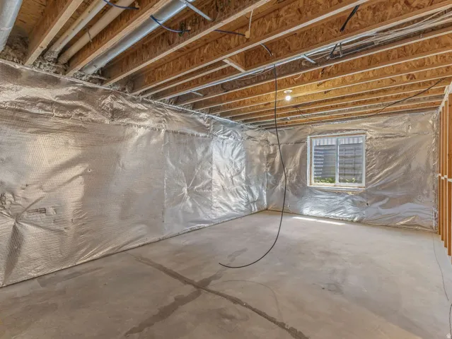 Unfinished basement space featuring a concrete floor, exposed wood ceiling joists, and a window with louvered blinds