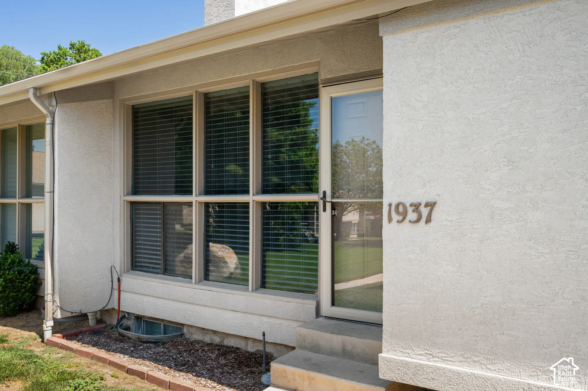 Stucco exterior featuring multiple large windows with blinds, a prominent address number, a front entry storm door, and a brick-edged landscape bed