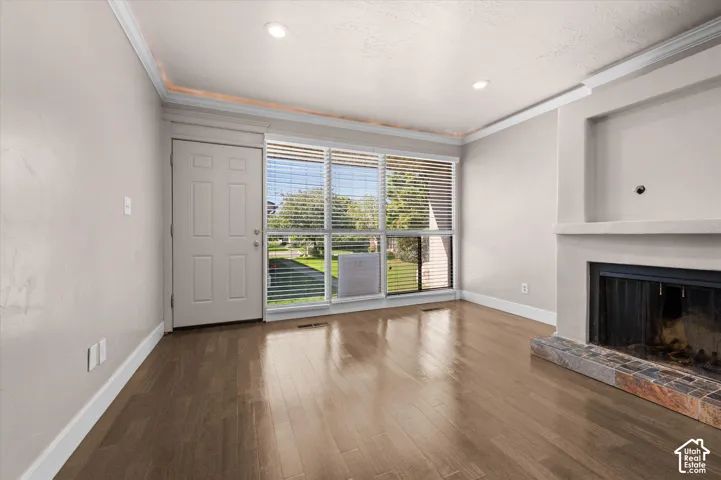 Warm wood-finish flooring, recessed lighting, crown molding, a fireplace with a stone hearth, and a large window with blinds