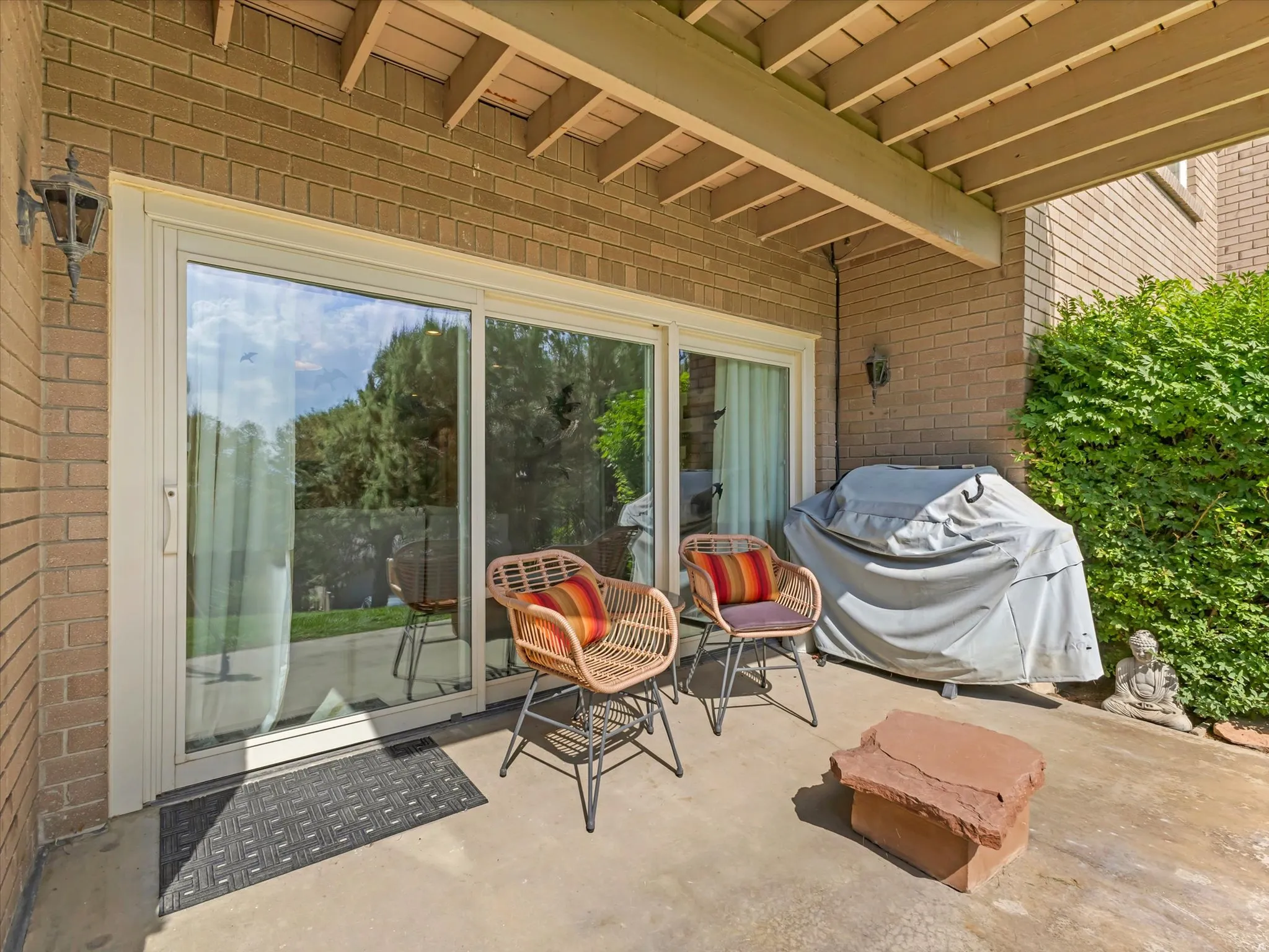 Patio area featuring brick exterior, concrete flooring, sliding glass door, overhead wooden beams, and mature landscaping