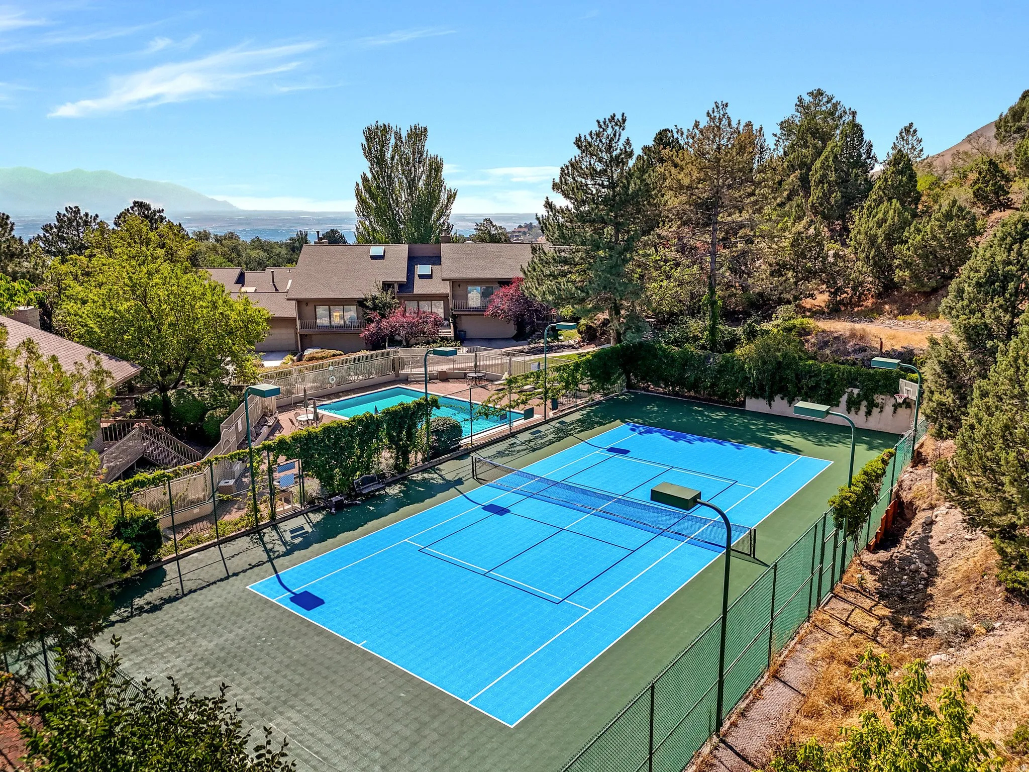 Blue-surfaced tennis court with perimeter fencing, outdoor swimming pool, and surrounding mature trees