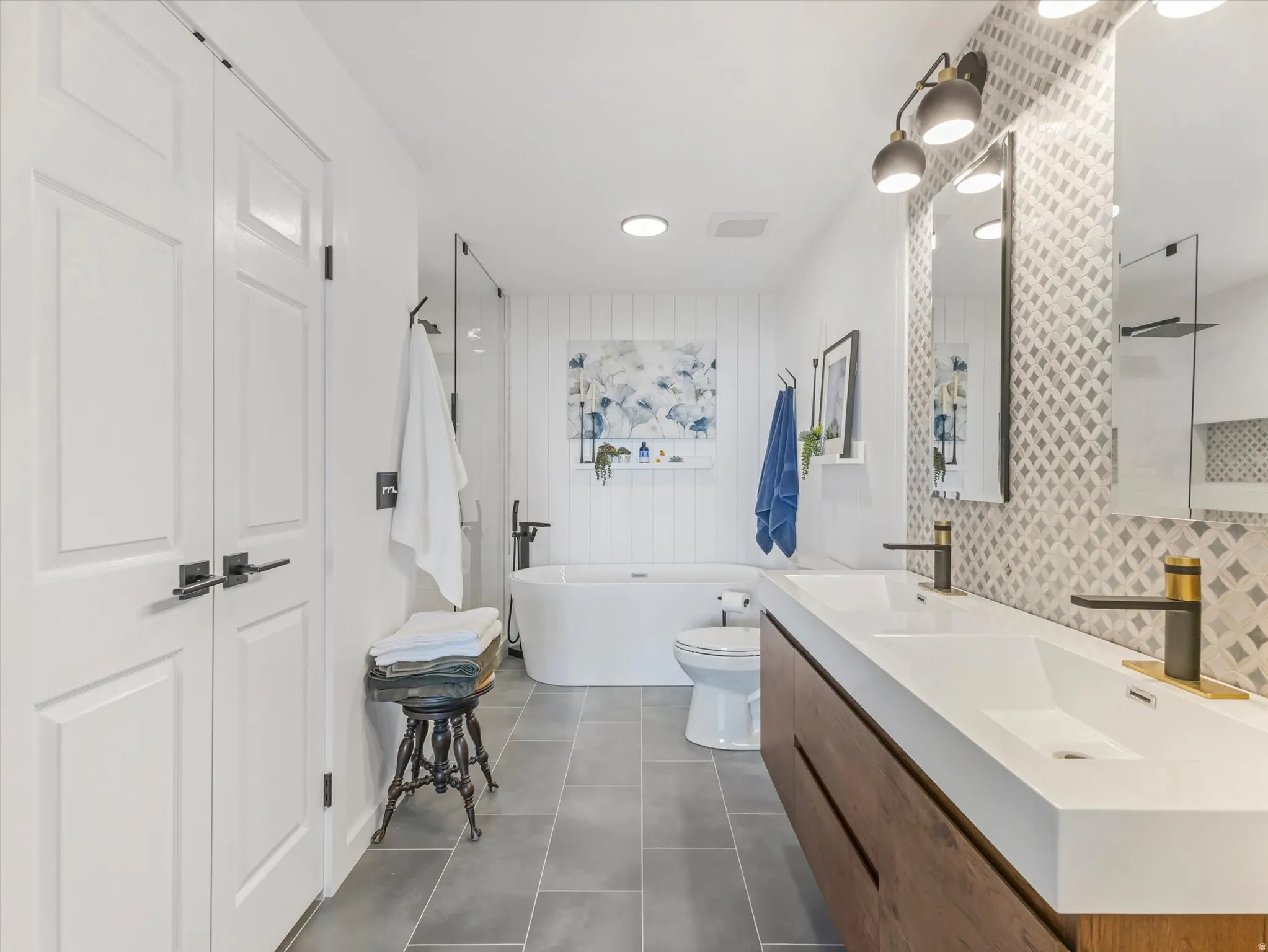 Contemporary bathroom featuring a floating wood-finish vanity with dual integrated sinks, striking geometric tile backsplash, and a freestanding soaking tub