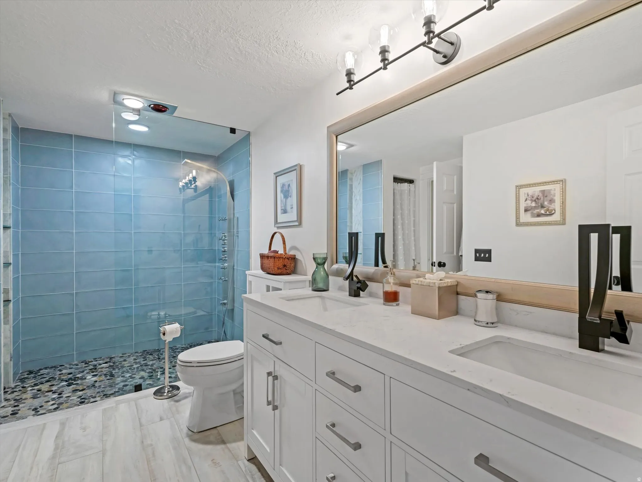 Bathroom featuring a double vanity with stone countertops and integrated sinks, a wide mirror with a wooden frame, and a frameless glass shower enclosure with blue tile surround and pebble flooring