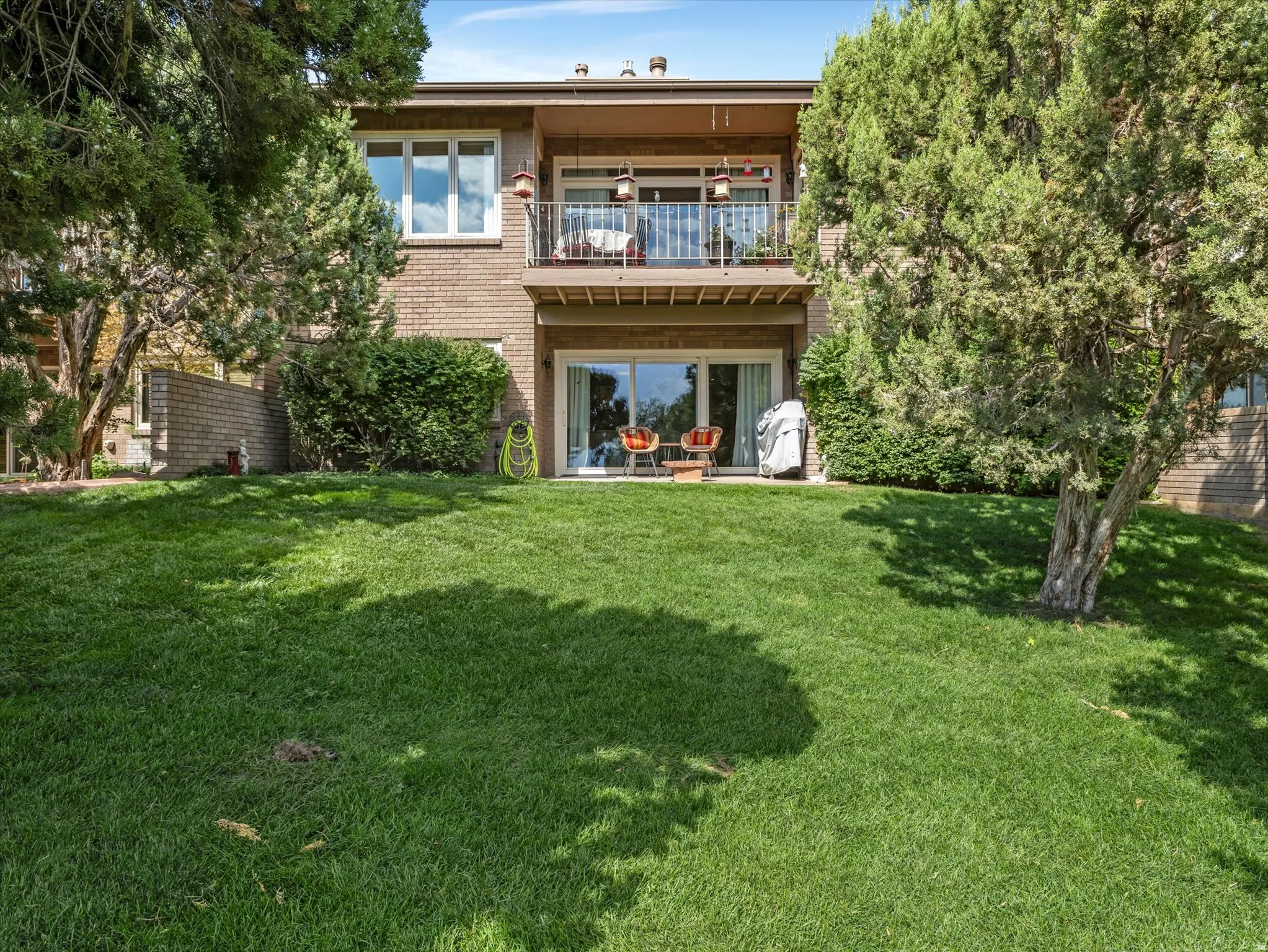 Rear exterior featuring a spacious lawn, mature trees, a ground-level patio with sliding glass doors, and an upper-level balcony with metal railings