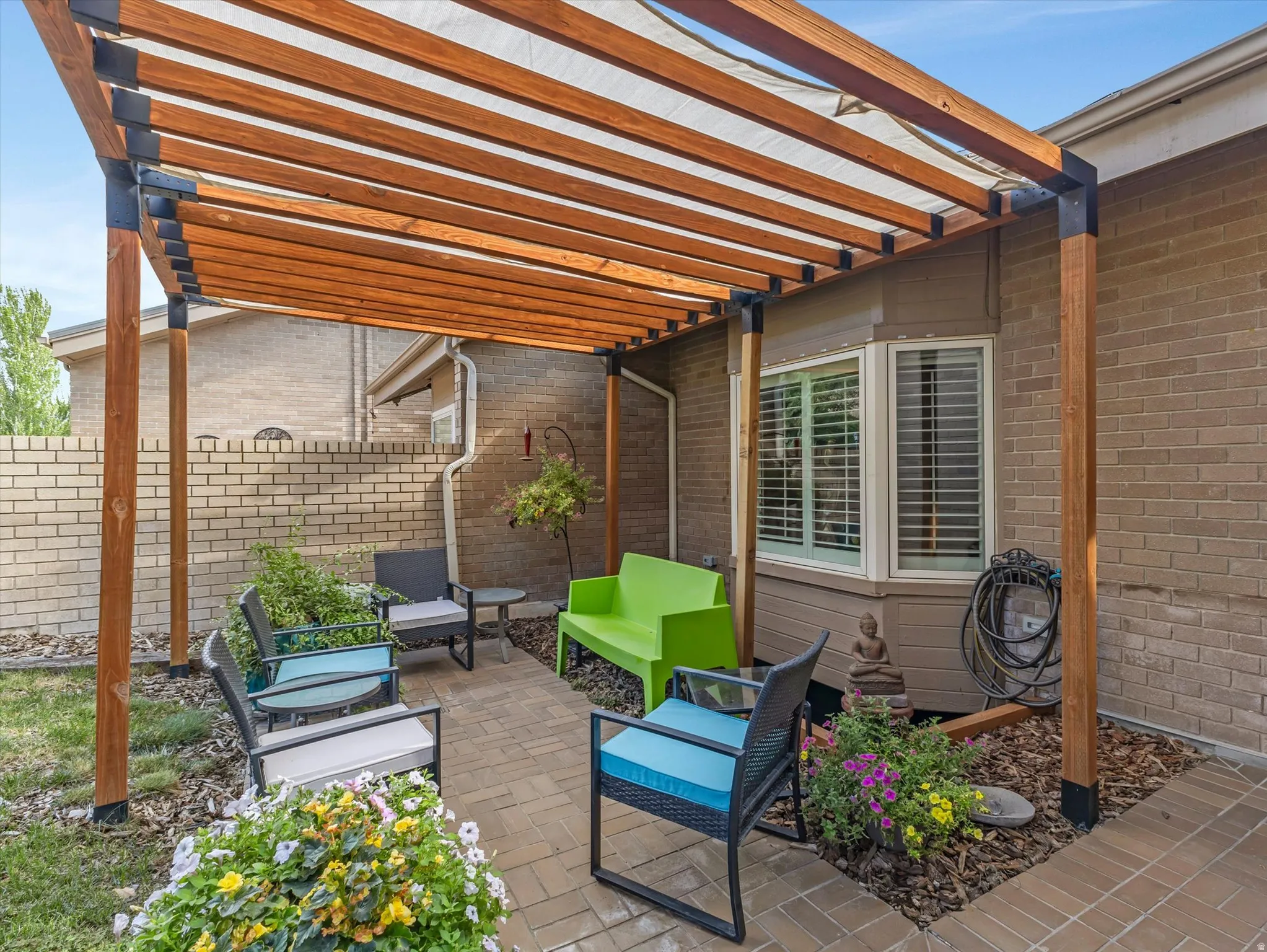 Outdoor patio featuring a wood pergola with a white fabric shade, brick paver flooring, and planted garden beds