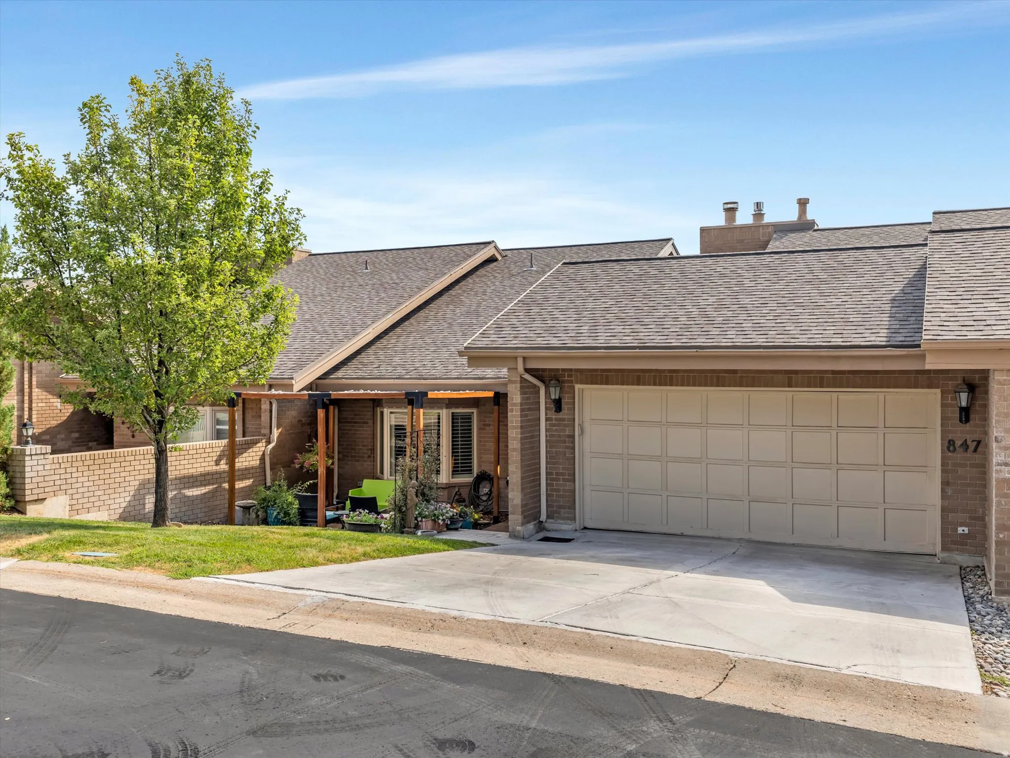 Brick-clad residential exterior featuring a two-car garage with a panel door, concrete driveway, and covered patio area with wood pergola