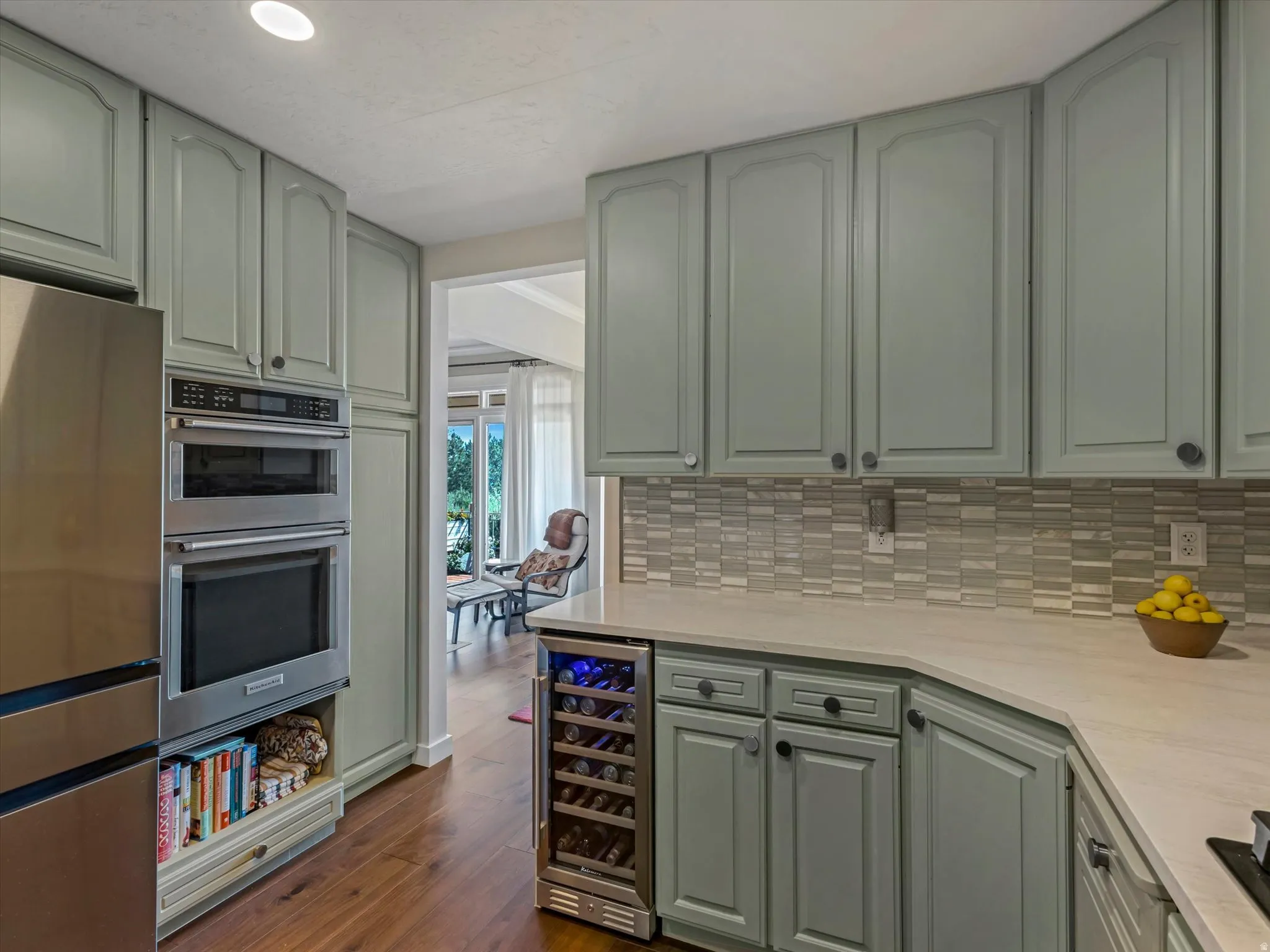 Kitchen featuring light green cabinetry, stainless steel appliances, a built-in wine refrigerator, a mosaic tile backsplash, and light-toned countertops