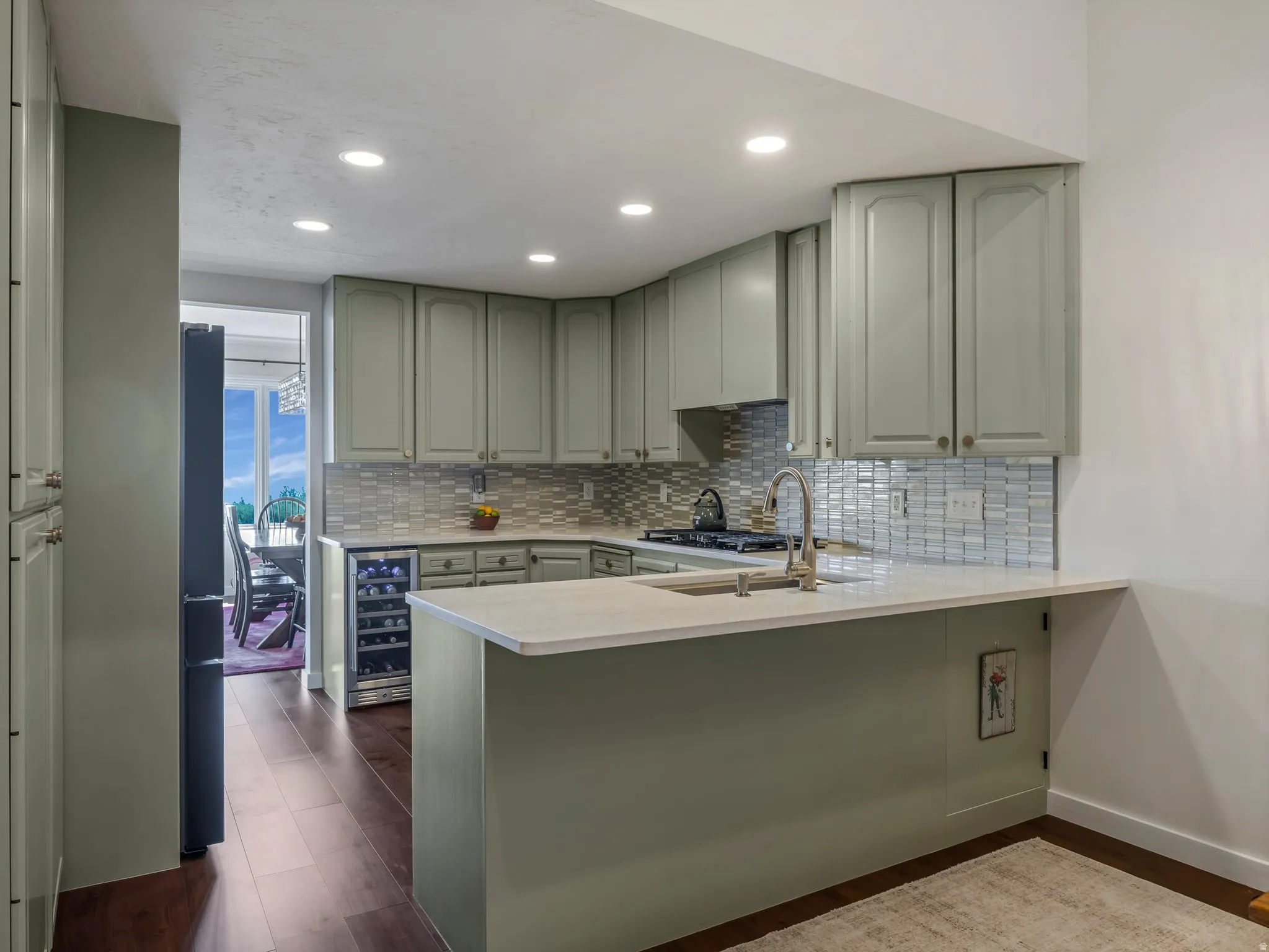 U-shaped kitchen with light green cabinetry, integrated wine cooler, light-toned countertops, tile backsplash, and dark wood-finish flooring