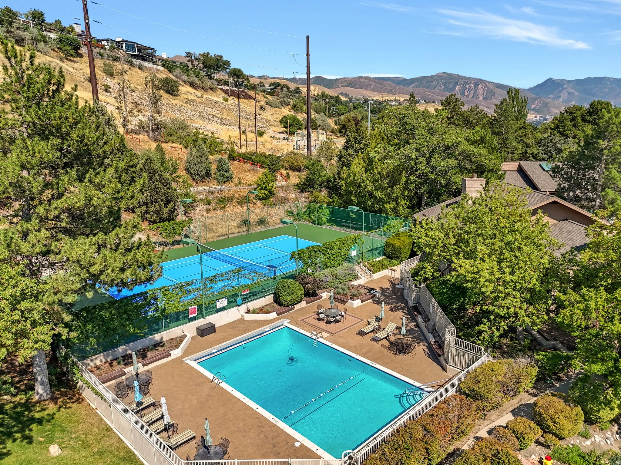 Outdoor pool with surrounding hardscaping, fenced tennis court, and lush mature landscaping