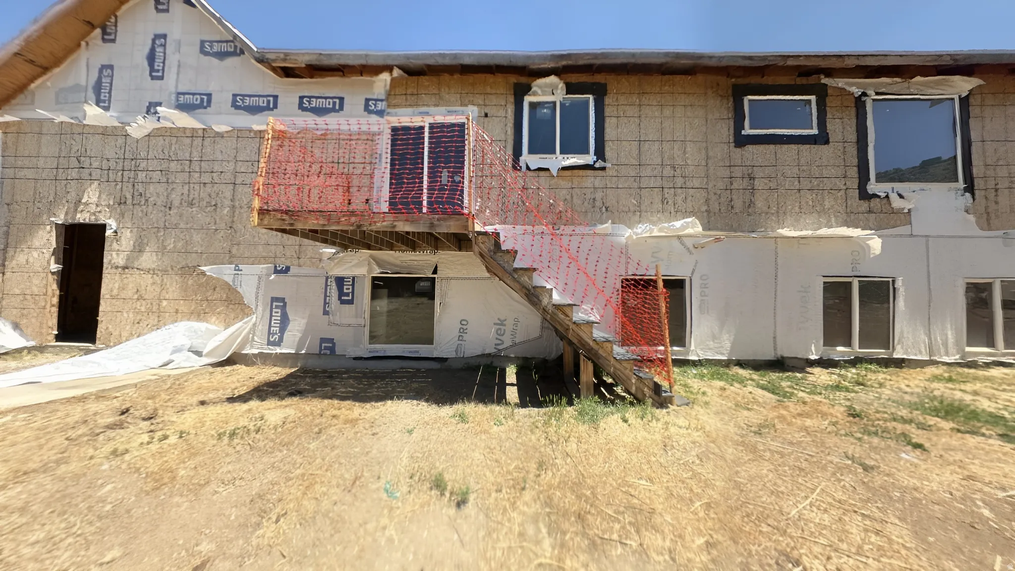 Exterior structure with visible sheathing and house wrap, featuring multiple window openings and a second-story wooden deck with stairs