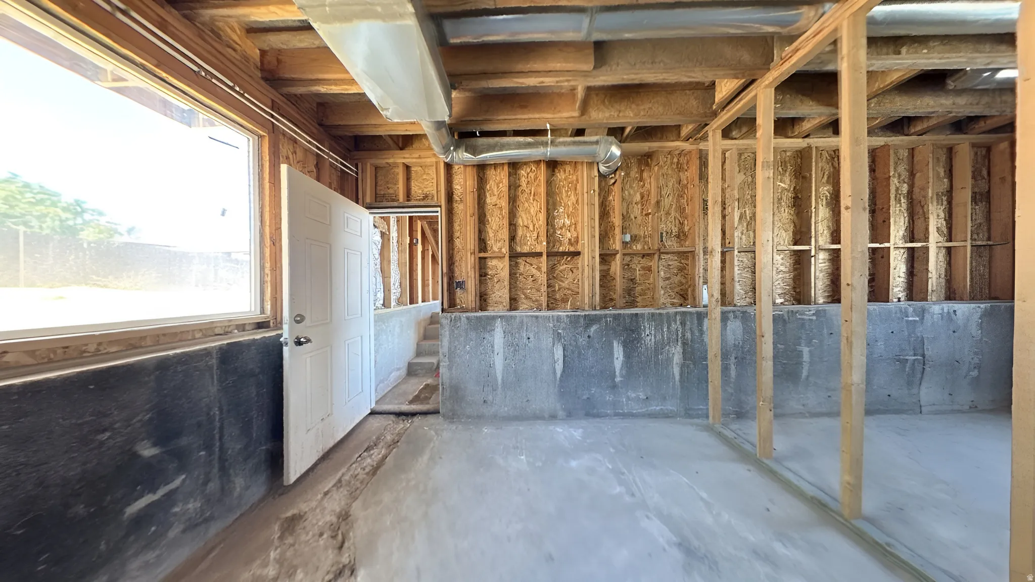 Unfinished interior space featuring exposed wood framing, concrete foundation walls, a large window, and an interior door