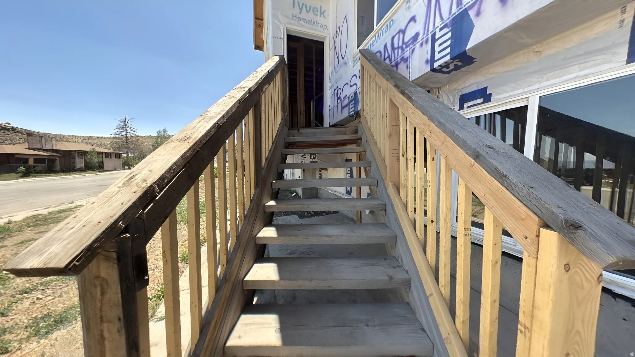 Exterior wooden staircase with baluster railing leading to a building under construction, featuring exposed Tyvek Homewrap and large reflective window panes