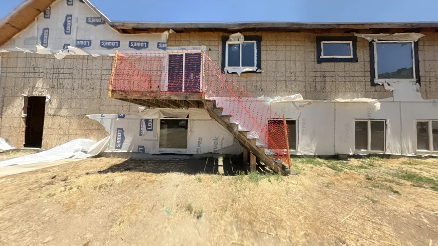 Exterior structure with visible sheathing and house wrap, featuring multiple window openings and a second-story wooden deck with stairs