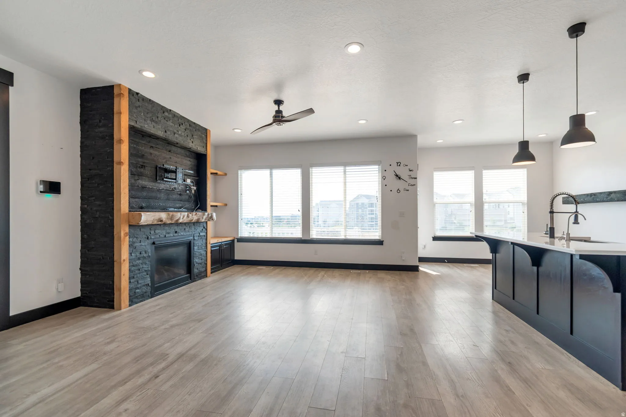 Spacious living area featuring light wood-finish flooring, a contemporary fireplace with a wood mantle and black brick surround, recessed lighting, and a ceiling fan