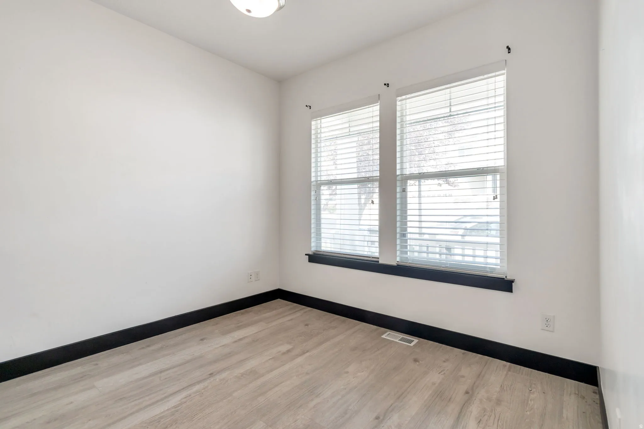Bright room featuring two windows with blinds, a flush mount ceiling light, light wood-finish flooring, and dark baseboards