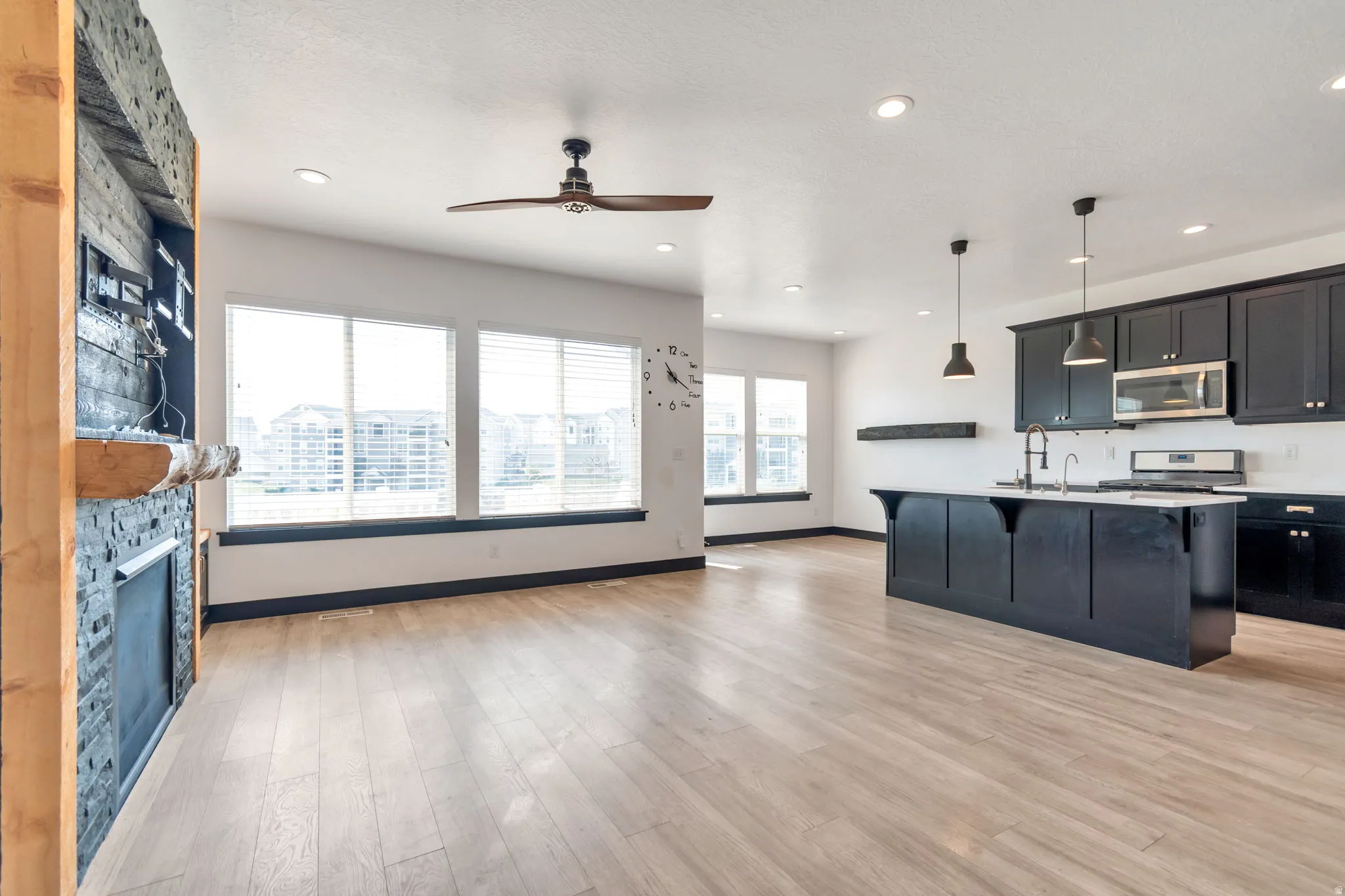 Open-concept living space featuring wood-finish flooring, a contemporary kitchen island with pendant lighting, black cabinetry, and stainless steel appliances