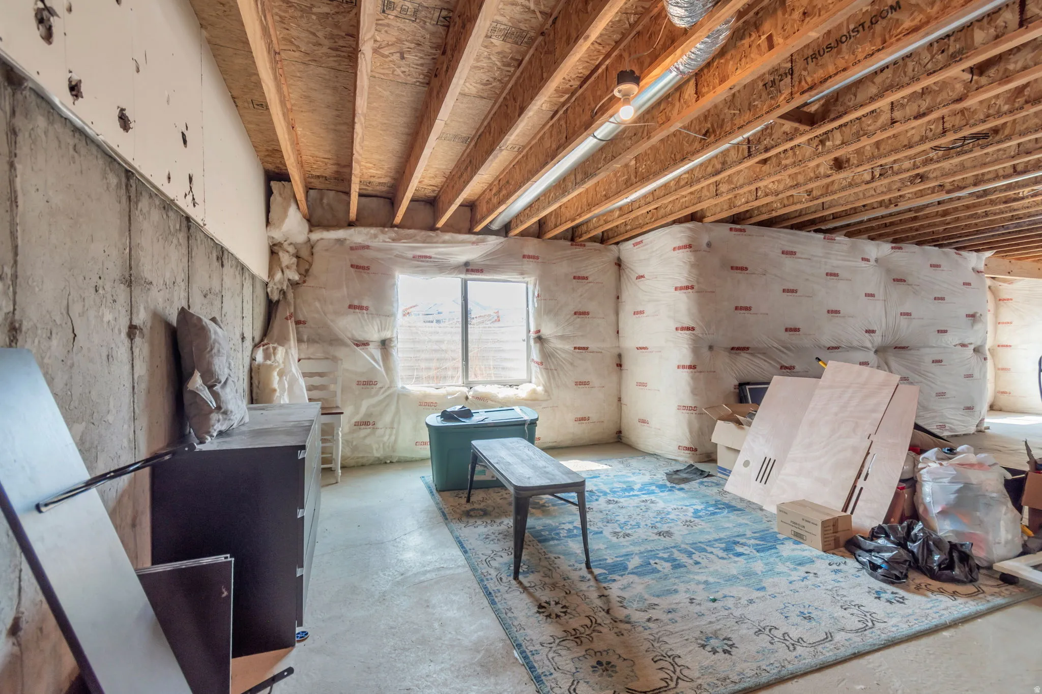 Unfinished basement space with exposed wooden ceiling joists, concrete flooring, and a visible window opening