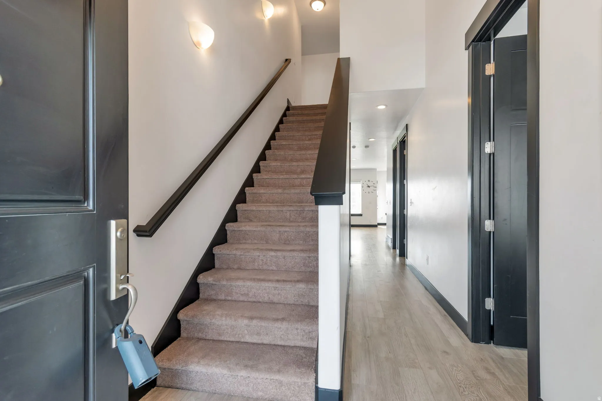 Inviting entryway featuring a dark paneled door with brushed nickel hardware, carpeted staircase with dark handrail and trim, and light wood-finish flooring extending into a hallway with recessed lighting and dark-trimmed doorways