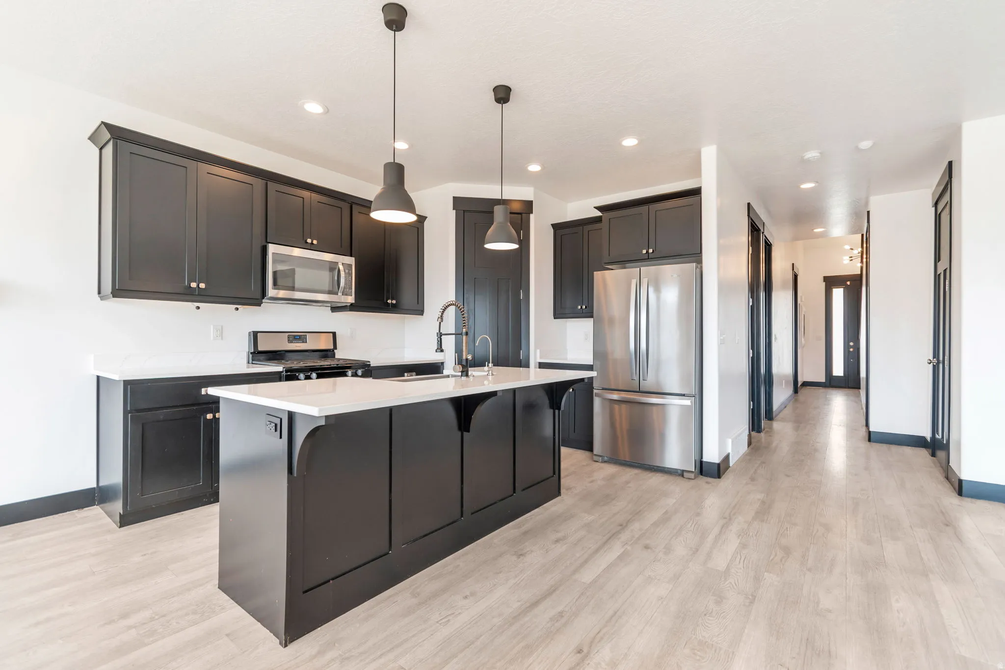 Modern kitchen featuring a large central island with a sink, dark cabinetry, stainless steel appliances, and light wood-finish flooring
