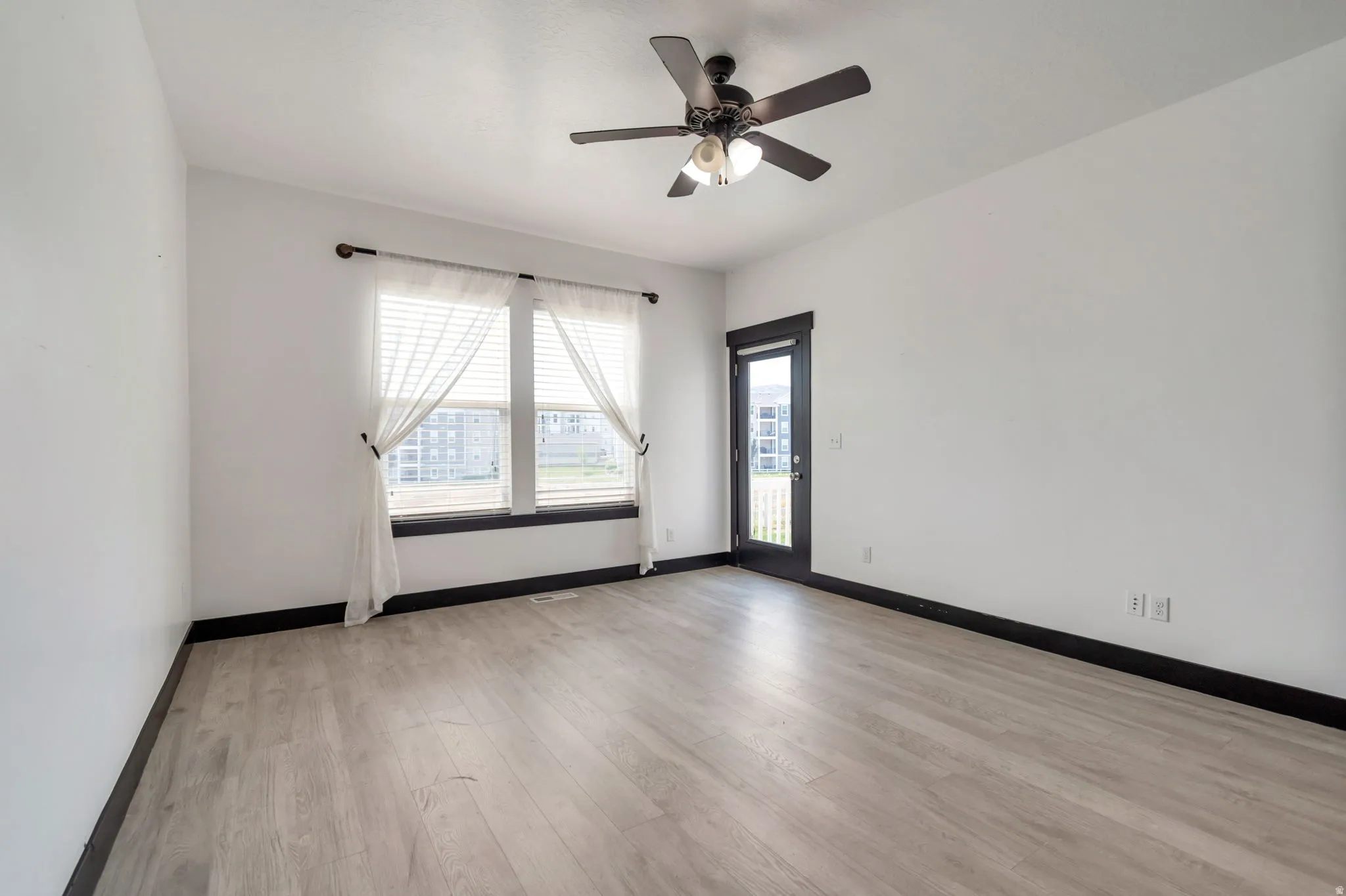 Spacious room featuring wood-finish flooring, a ceiling fan with integrated lighting, and a large window with dark trim