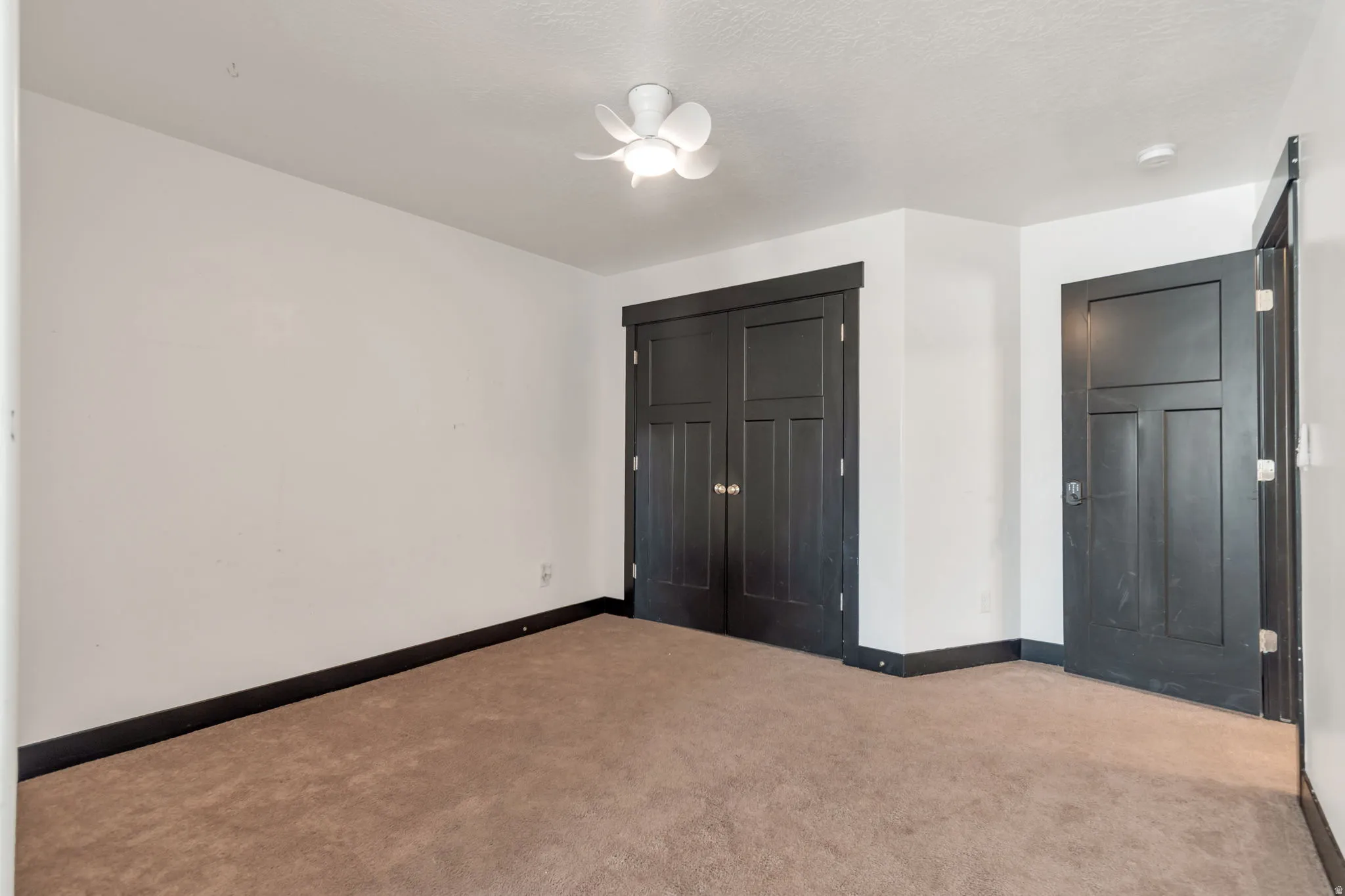 Carpeted room featuring a ceiling fan with light fixture, white walls, dark trim, a dark paneled door, and dark bi-fold closet doors